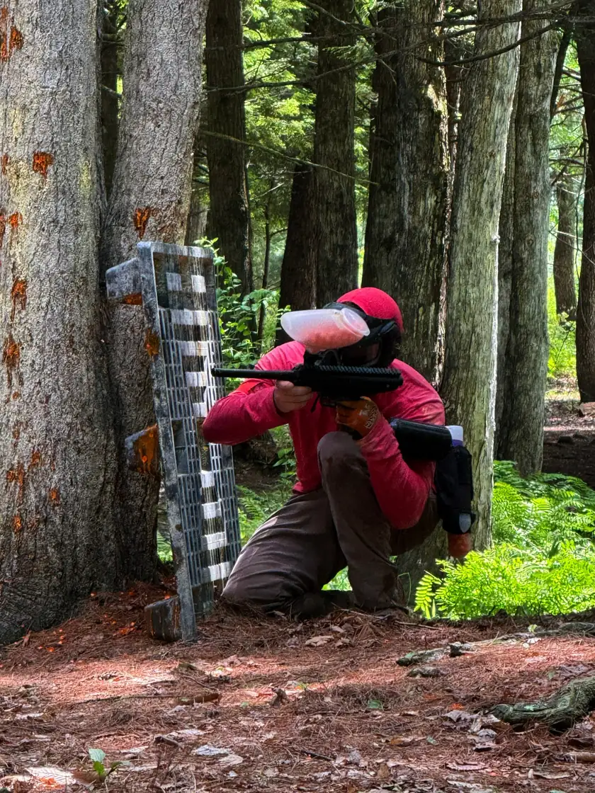 Player in red kneeling behind a bunker against a tree, aiming through the woods
